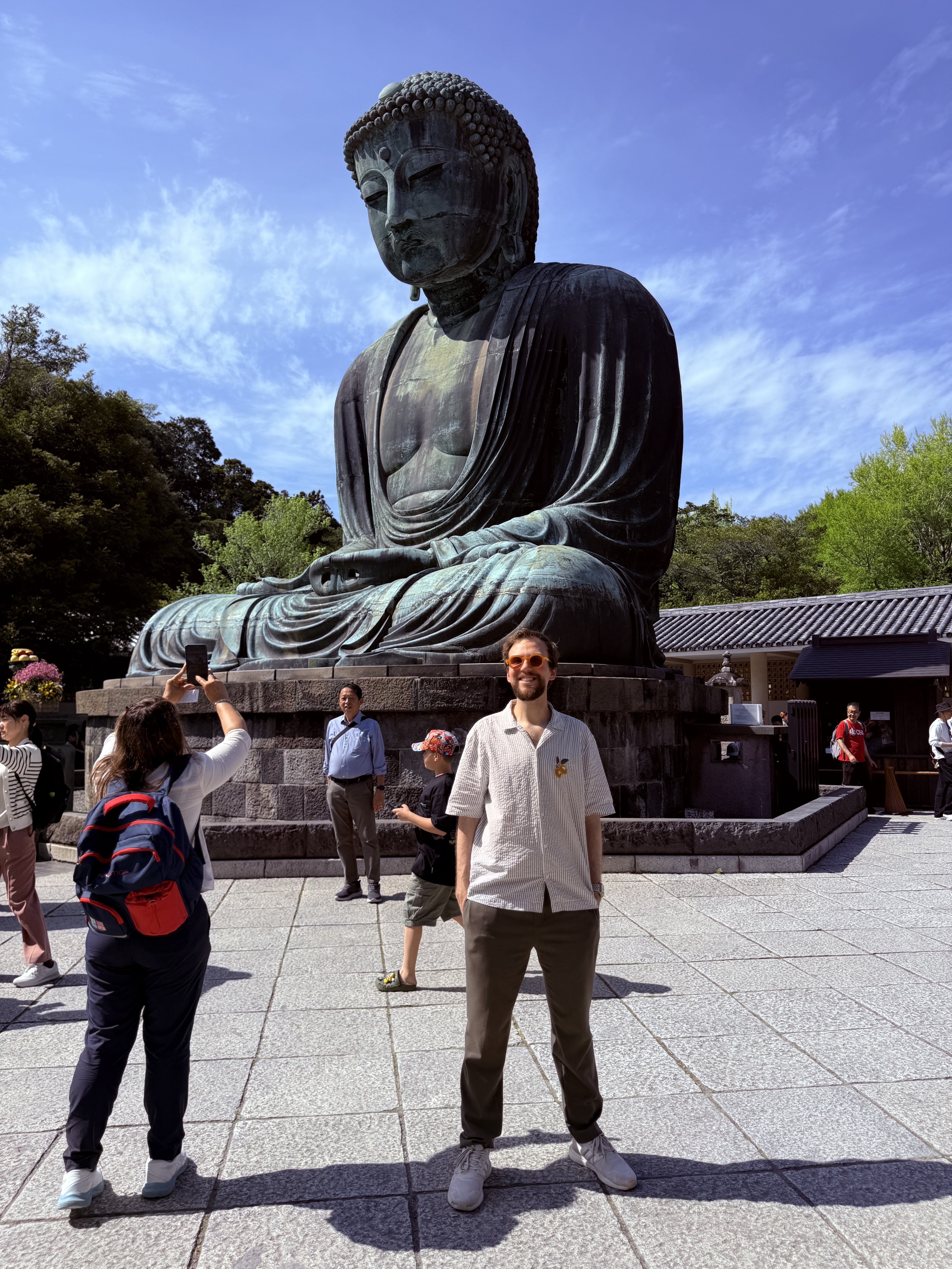 Me posing and smiling in front of the Great Buddha (Daibutsu) in Kamakura on a sunny day, wearing sunglasses and a light striped shirt, with other tourists visible in the background.