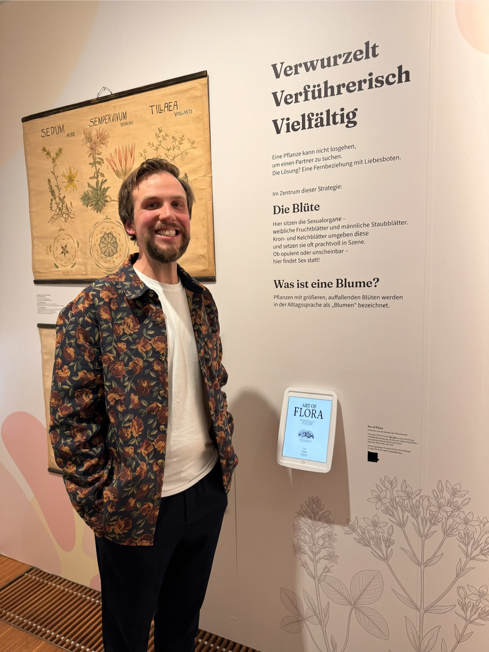 Me in a floral jacket standing next to an iPad displaying Art of Flora at the BLOOM 'Blümchensex' exhibition, beside a vintage botanical teaching chart of Sedum, Sempervivum, and Tillaea, with German exhibition text about plant reproduction on the wall.