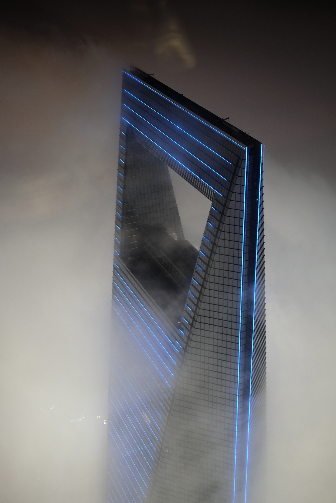 The top of the Shanghai World Financial Center at night, its distinctive trapezoidal aperture glowing with blue LED light strips, emerging from thick fog.