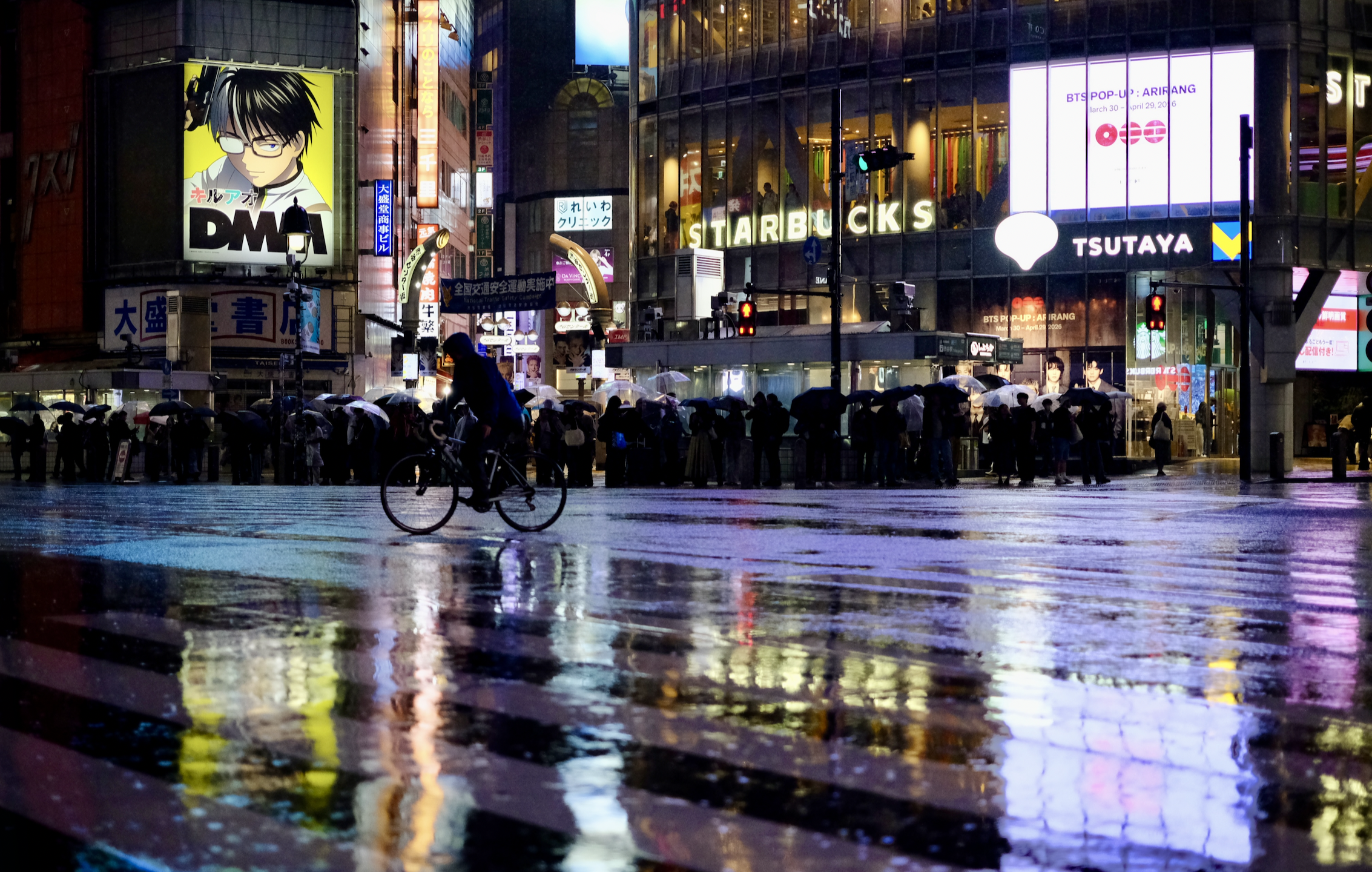 Shibuya Crossing on a rainy night — a lone cyclist rides across the wet pavement, neon signs and storefronts (Starbucks, Tsutaya) reflecting in the rain-soaked street, with crowds of umbrella-holding pedestrians waiting at the curb.